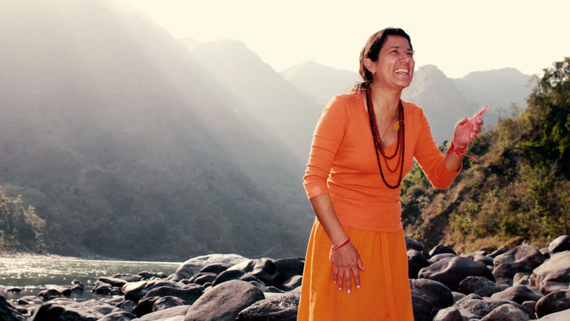 Mataji Shaktiananda in orange, laughing on the rocky bank of a mountain river